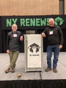 Two gentlemen standing either side of a podium on a stage, with a green and black sign behind them that reads NY RENEWS.