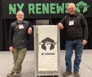 Two gentlemen standing either side of a podium on a stage, with a green and black sign behind them that reads NY RENEWS.