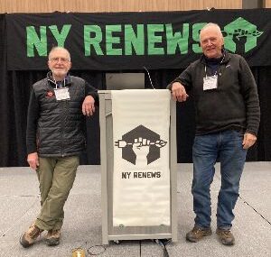Two gentlemen standing either side of a podium on a stage, with a green and black sign behind them that reads NY RENEWS.